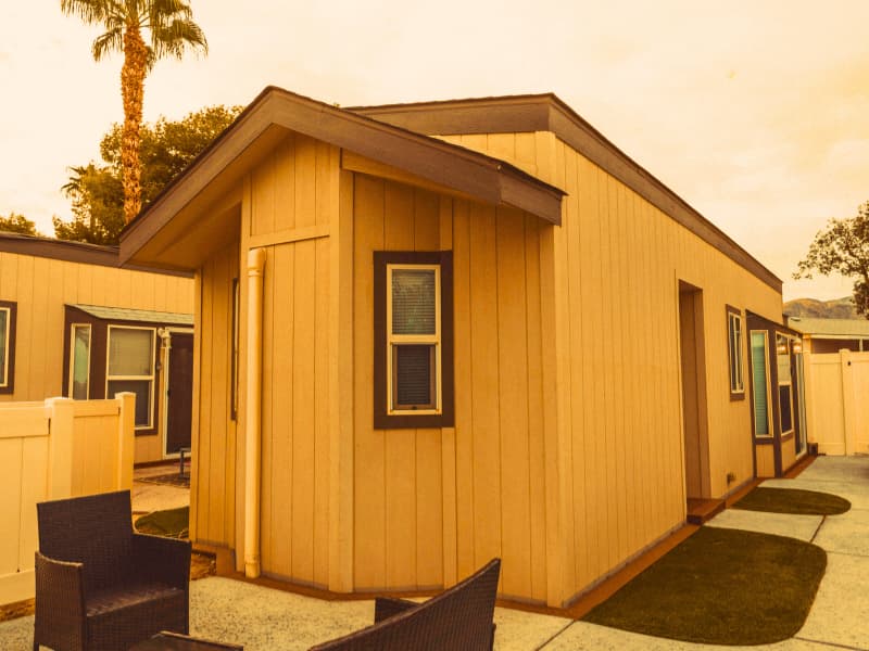Compact modular home with vertical wood siding and a small patio area, surrounded by a privacy fence and palm trees.