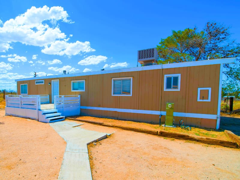 Traditional mobile home with beige exterior, white trim, and front porch access via a small ramp, located in a dry rural setting.