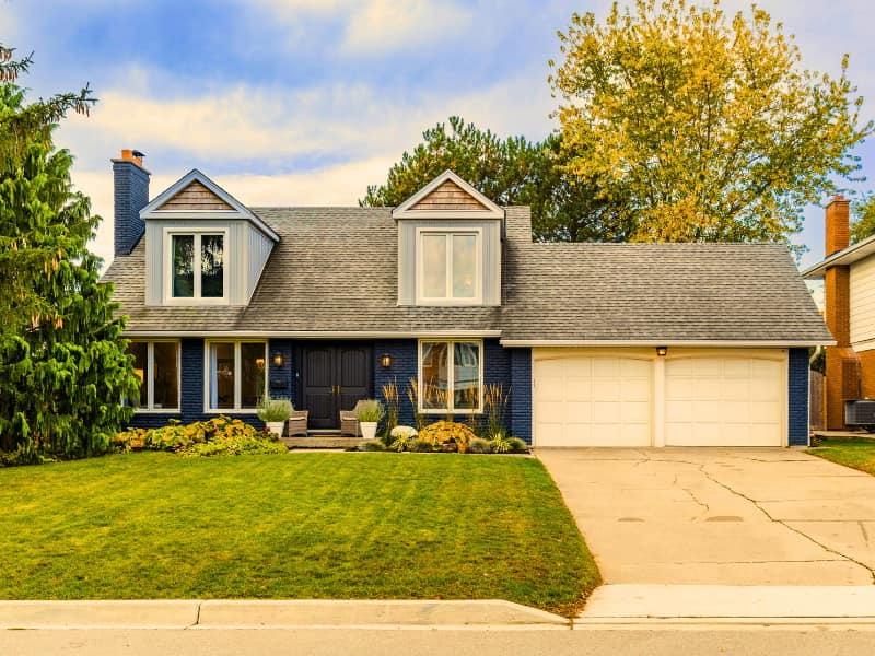 Two-story blue home with dormer windows, landscaped front yard, and mature trees — an example of how exterior upkeep and surroundings affect the best home insurance choices.