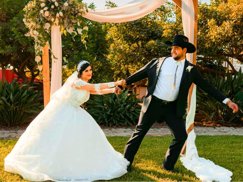 Bride playfully pulling the groom in a cowboy hat beneath a draped outdoor arch—garden celebration that benefits from wedding venue insurance.
